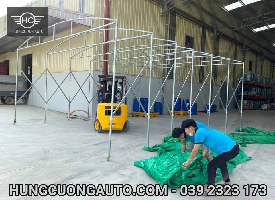Hai người chuẩn bị bạt xanh bên khung mái vòm và xe nâng trước xưởng