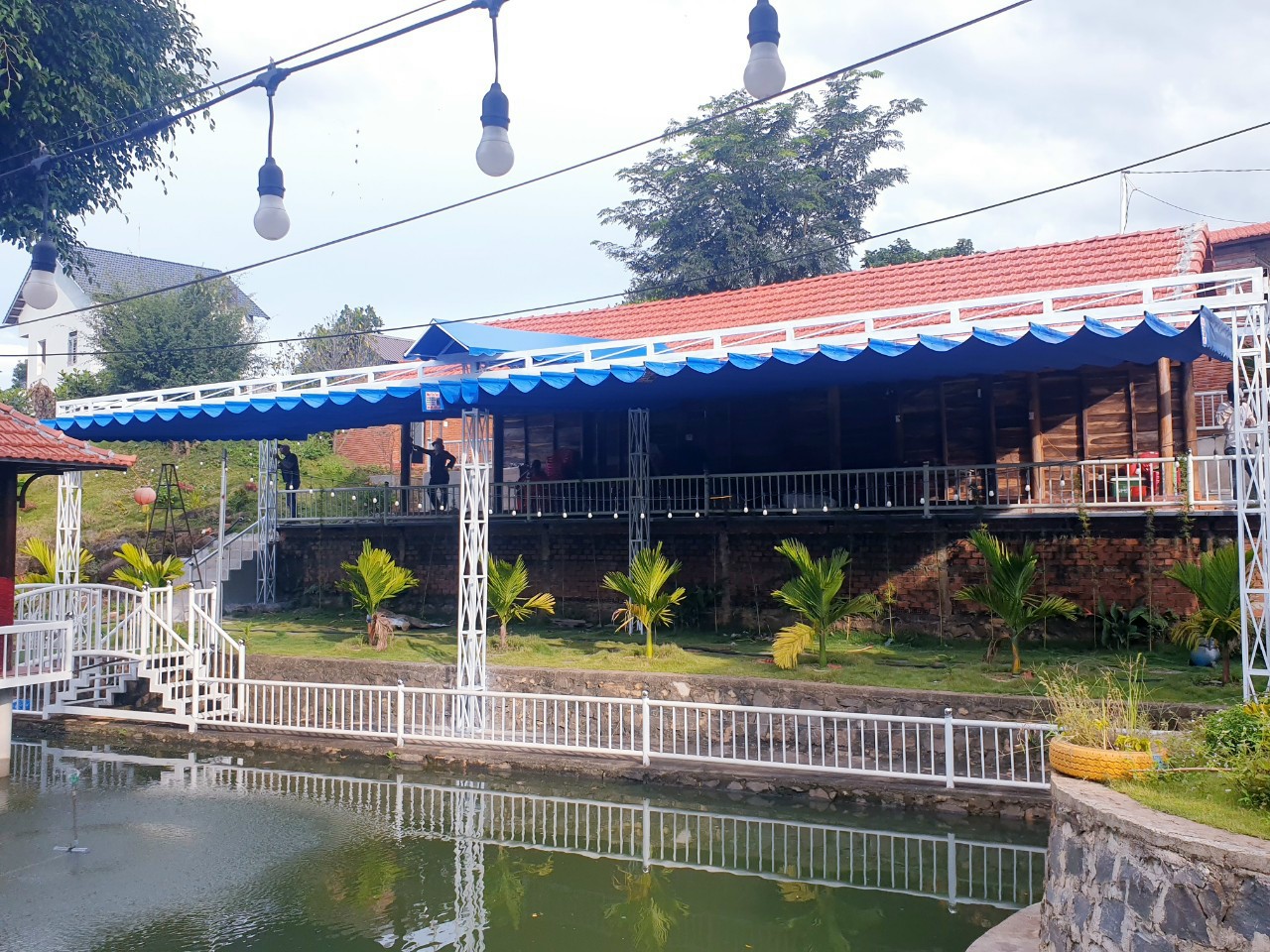 Mái bạt xanh phía trước nhà, cạnh hồ nước có lan can và cây cảnh